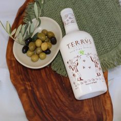 Botella de aceite de oliva virgen extra Terrvs junto a un plato con aceitunas verdes y negras sobre una tabla de madera.